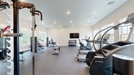 Located at 562 Centennial Pkwy in Tyler, TX, this fitness center offers a well-appointed exercise space designed to support a full range of workout routines. The single-floor layout is bright and open, featuring crisp white walls, recessed lighting, and durable rubber flooring that provides both safety and comfort underfoot.

The cardio zone is anchored by multiple treadmills, an elliptical trainer, a stair climber, and a rowing machine, all positioned along a windowed wall that fills the space with natural light and views of surrounding greenery. A dedicated strength training area houses a multi-functional cable machine and a full power rack with weight plates, offering a comprehensive setup for resistance training.

Free weights are neatly organized on a tiered dumbbell rack, accompanied by kettlebells and a flat utility bench, providing versatility for a variety of exercises. A wall-mounted flat-screen television enhances the workout experience. Additional accessories, including medicine balls and resistance equipment, are stored on nearby shelving. The multiple windows on adjacent walls bring in ample daylight while maintaining a clean, energizing atmosphere. The space is thoughtfully arranged to maximize functionality while allowing comfortable movement throughout, making it a practical and fully equipped fitness facility.