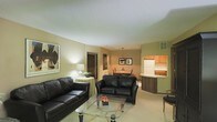 This single-floor space offers an inviting open-concept layout that seamlessly connects the living, dining, and kitchen areas into one cohesive and functional environment. The living area is anchored by a pair of dark leather sofas arranged around a glass-topped coffee table, creating a comfortable and stylish seating arrangement. Warm neutral tones coat the walls throughout, complemented by soft lighting from a classic table lamp and overhead pendant fixtures that enhance the welcoming ambiance. A framed piece of abstract wall art adds a touch of character to the living space. The dining area transitions naturally from the living room and features a round table with wooden chairs, positioned beneath a pendant light and accented by a small floral centerpiece. Just beyond, the kitchen is defined by a curved breakfast bar with warm wood cabinetry, offering additional casual seating and workspace. The kitchen is equipped with standard appliances, including a full-size refrigerator, and features upper cabinetry providing ample storage. Light-toned flooring runs throughout the space, tying the areas together and contributing to a bright, airy feel. The overall design is practical and comfortable, making the space well-suited for both relaxing and everyday living.