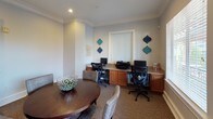 This single-floor space combines a comfortable meeting area with a functional business center, making it an ideal environment for both collaborative gatherings and individual productivity. The meeting area is anchored by a rich, dark-stained round wooden table surrounded by upholstered gray chairs, creating an inviting setting for small group discussions or casual dining. Built-in open shelving units flank the walls, offering tasteful display space accented with decorative objects and framed abstract artwork in teal and navy tones. Elegant crown molding runs throughout the space, lending a polished, traditional character to the soft gray walls and neutral carpeted floors. Natural light filters in through window blinds, brightening the interior. A classic wood-framed exterior door with sidelights provides direct outdoor access. French doors with frosted glass panes connect the meeting area to adjacent spaces, maintaining a sense of openness while offering separation. The business center area features a built-in wood-cabinet workstation with granite-style countertops, housing dual computer workstations equipped with monitors, keyboards, and a shared printer — ideal for focused work sessions. Decorative geometric wall accents in teal and navy add a contemporary touch, tying the overall aesthetic together cohesively.