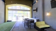 This single-floor amenity lounge blends indoor comfort with an open, airy atmosphere. A striking floor-to-ceiling glass wall floods the space with natural light while offering views of the surrounding outdoor area, which features artificial turf and a gated patio beyond. The interior is defined by polished tile flooring and charcoal-toned walls that lend a modern, urban aesthetic. A built-in bar counter with a beverage tap runs along one wall, accompanied by high-top tables and wicker bar stools that encourage social gathering. A wall-mounted flat-screen TV is positioned nearby, making the space suitable for casual viewing. Warm string lights are strung across the ceiling, adding a relaxed, inviting ambiance. A wicker sofa with bright yellow cushions provides a comfortable lounge seating option, introducing a pop of color that complements the modern palette. The arched architectural detail framing the glass wall adds a subtle design flourish, while the overall layout balances communal gathering and casual relaxation in an open, well-lit environment.