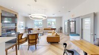 Located on Park Lane in Dunellen, NJ, this thoughtfully designed single-floor amenity space offers residents a comprehensive and modern environment for both relaxation and activity. The open-concept lounge and kitchen area serves as the heart of the space, featuring a striking herringbone-patterned hardwood floor, a large white quartz island with bar seating, warm wood cabinetry, stainless steel appliances, and globe pendant lighting that lends a contemporary sophistication. A mounted TV is set against a textured stone accent wall, while comfortable seating arrangements invite casual socializing. The lounge area is anchored by a bold circular chandelier and furnished with a mix of leather and upholstered seating, complemented by ample natural light streaming through large windows that frame views of the surrounding community. The expansive fitness center is outfitted with a full suite of cardio and strength equipment, including treadmills, stationary bikes, ellipticals, and free weights, all set beneath bright panel lighting with wall-to-wall mirrors and large windows providing natural light and outdoor views. A versatile flex room with soft carpet flooring and arched windows offers additional functionality, while well-connected hallways ensure smooth circulation throughout the space.