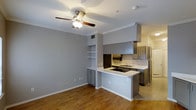 This single-floor Dallas space presents an open-concept design that seamlessly connects the living area and kitchen. The living space features warm hardwood flooring, crown molding, and a ceiling fan that enhances both comfort and airflow. Built-in shelving with cabinetry is integrated into the wall, offering functional storage while adding architectural character to the room.

The kitchen is defined by a wraparound peninsula with a light-colored countertop that provides both prep space and a casual dining option. Gray cabinetry extends throughout, complementing the neutral gray wall tones for a cohesive, modern aesthetic. Stainless steel appliances, including a full-size refrigerator, are incorporated into the kitchen layout. An overhead cabinet structure frames the cooking area, adding visual depth.

The transition between the living area and kitchen is smooth, with the peninsula acting as a natural divider without closing off the space. Tile flooring in the kitchen contrasts with the hardwood in the living area, subtly distinguishing each zone. Overall, the space offers a clean, contemporary atmosphere with practical design elements throughout.
