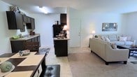 This single-floor residence showcases an open-concept floor plan that integrates the kitchen, dining, and living spaces into one cohesive and functional layout. The kitchen is outfitted with dark espresso cabinetry that creates a bold contrast against light-colored walls, and includes a black refrigerator, ample counter space with light-toned countertops, and additional tall cabinetry for generous storage. A peninsula-style counter serves as a natural transition point between the kitchen and the adjacent dining area, which features a dark dining table paired with leather-accented chairs. The flooring transitions from tile in the kitchen and dining zone to plush carpeting in the living area, adding warmth and comfort underfoot. The living room is spacious and inviting, anchored by a neutral-toned sofa and accented with soft lamp lighting that contributes to a cozy ambiance. Clean white walls and a simple, uncluttered aesthetic keep the interior feeling bright and open throughout. A white interior door provides access to additional areas of the home. The overall design balances practicality with a contemporary style, making it a comfortable and livable space.
