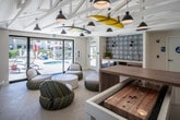 Located at 260 Avenida Vista Montana in San Clemente, California, this single-floor community amenity space captures the laid-back spirit of Southern California's surf culture. The room's most striking design element is the collection of colorful surfboards — including a vivid yellow and a multicolored board — suspended horizontally from the exposed white beam ceiling, instantly setting a coastal tone. Matte black pendant lights hang throughout, adding a modern contrast to the bright, airy interior.

The lounge area features round woven accent chairs with plush white cushions arranged around a terrazzo-topped coffee table, creating an inviting, casual gathering space. A bold black-and-white patterned tile accent wall serves as a focal point, complemented by warm wood cabinetry with under-counter storage below. A built-in wooden counter along one wall is paired with modern white bar stools, offering a workspace or casual dining perch beneath colorful framed artwork.

A large sliding glass door floods the space with natural light and opens directly to an outdoor pool deck, where lounge chairs and umbrellas are visible, seamlessly blending indoor and outdoor living. The light-toned flooring and crisp white walls enhance the sense of brightness and openness throughout, making this a vibrant and functional communal retreat.