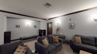 This theater lounge is a thoughtfully designed single-floor space that combines comfort and functionality in a warm, inviting setting. The room is finished with light blue-gray walls complemented by rich dark wood crown molding and baseboards, creating a refined yet relaxed atmosphere throughout.

Seating is a central focus of the space, with multiple dark charcoal sofas and accent chairs arranged to encourage both conversation and viewing. Colorful chevron-patterned throw pillows add visual interest and a touch of personality to the neutral furnishings. A woven upholstered bench and additional lounge chairs round out the flexible seating arrangements.

At one end of the room, a large wall-mounted flat-screen television serves as the entertainment focal point, positioned above a dark curved media console. Wall-mounted sconce lighting is distributed throughout, providing warm, even illumination across the carpeted space.

A bookcase stocked with reading materials is tucked into one corner, while a console table with decorative accessories adds an element of style. Framed black-and-white photographs adorn the walls, lending a curated, gallery-like quality to the room. A high-top dining table with chairs offers an additional seating option for those preferring an upright position.

Double dark wood doors provide access to the space, reinforcing the cohesive, classic design aesthetic present throughout this versatile theater lounge.