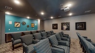 This private screening room is designed to deliver an immersive cinematic experience. The room is arranged with multiple rows of plush, blue upholstered armchairs positioned in a tiered layout facing a large wall-mounted projection screen. A ceiling-mounted projector provides high-quality image display, while recessed lighting in the dark-painted ceiling creates a theater-like ambiance that enhances viewing comfort.

The seating arrangement accommodates a generous number of viewers, with chairs organized in clearly defined rows atop a patterned, neutral-toned carpet. The muted gray walls are complemented by a vibrant teal accent wall at the rear of the room, which serves as a backdrop for decorative movie-themed artwork, including film reel motifs and popcorn signage that reinforce the entertainment atmosphere.

A built-in snack bar along the teal accent wall features a countertop with a sink, adding a convenient concession-style amenity to the space. Additional framed cinema-themed artwork adorns the side walls, further enhancing the theatrical character of the room. The overall design balances functional layout with thoughtful décor, creating a dedicated, well-appointed venue for group screenings and entertainment.