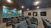 This private screening room is designed to deliver an immersive cinematic experience. The room is arranged with multiple rows of plush, blue upholstered armchairs positioned in a tiered layout facing a large wall-mounted projection screen. A ceiling-mounted projector provides high-quality image display, while recessed lighting in the dark-painted ceiling creates a theater-like ambiance that enhances viewing comfort.

The seating arrangement accommodates a generous number of viewers, with chairs organized in clearly defined rows atop a patterned, neutral-toned carpet. The muted gray walls are complemented by a vibrant teal accent wall at the rear of the room, which serves as a backdrop for decorative movie-themed artwork, including film reel motifs and popcorn signage that reinforce the entertainment atmosphere.

A built-in snack bar along the teal accent wall features a countertop with a sink, adding a convenient concession-style amenity to the space. Additional framed cinema-themed artwork adorns the side walls, further enhancing the theatrical character of the room. The overall design balances functional layout with thoughtful décor, creating a dedicated, well-appointed venue for group screenings and entertainment.