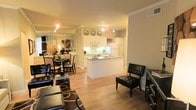 This single-story space in Irving, Texas, showcases an open-concept design that connects the living, dining, and kitchen areas into one cohesive, flowing layout. The kitchen is outfitted with white cabinetry extending to the ceiling, white appliances, and a peninsula with an integrated sink that serves as a natural dividing point between the kitchen and the surrounding living spaces. Under-cabinet lighting and track lighting throughout provide warm, even illumination across the space. Crown molding along the ceilings adds a refined architectural detail that elevates the overall aesthetic. The dining area, positioned adjacent to the kitchen, features a dark-toned table and chair set, complemented by decorative accents including metallic wall ornaments that add visual interest. A large mirror in the dining area enhances the sense of openness and reflects light throughout the room. The living area accommodates tufted leather seating and a patterned area rug, contributing to a polished, contemporary feel. Warm hardwood-style flooring runs consistently throughout, unifying the entire space. The overall ambiance is bright, well-appointed, and cohesive, making efficient use of the open floor plan.