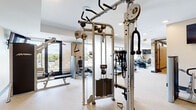 Located at 871 Sibley Memorial Hwy in Lilydale, MN, this comprehensive fitness center offers a thoughtfully designed, single-level layout spanning a generous floor area suited for a full range of exercise activities. The main gym floor is bright and open, bathed in natural light streaming through floor-to-ceiling windows that offer views of the outdoor pool area. This section houses an impressive collection of strength training equipment, including a multi-function cable machine, weight benches, selectorized machines, and a dumbbell rack, complemented by a wall-mounted flat-screen TV. A row of cardio machines — including treadmills, ellipticals, and a stair climber — lines the window-facing wall, allowing users to exercise with scenic outdoor views. A dedicated water station and built-in cabinetry add functional convenience. A separate studio room with light hardwood flooring, ceiling fans, and full-length mirrors provides an ideal space for cycling classes, barre workouts, and group fitness, with ICG spin bikes, stability balls, and resistance equipment readily available. Recessed lighting throughout maintains a clean, modern aesthetic, while the neutral warm-toned walls and speckled flooring create a polished, professional atmosphere across the entire facility.