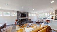 This single-floor space showcases a contemporary open-concept design, seamlessly blending comfort and modern aesthetics throughout. The centerpiece of the main living area is a striking floor-to-ceiling mosaic tile accent wall, framing both a built-in fireplace and a large flat-screen television. Recessed lighting illuminates the space, complementing the large-format tile flooring accented with hardwood inlays. The living area is generously proportioned, accommodating multiple seating arrangements including leather sofas and accent chairs, anchored by a bold floral-patterned area rug in warm amber tones. Adjacent to the living space, a stylish bar counter with pendant lighting and bar stools provides an ideal casual gathering spot. The open layout flows naturally into a dining area, which is bathed in natural light from a sweeping bank of floor-to-ceiling windows and a glass door that opens onto an outdoor deck. The deck overlooks a lush, tree-lined landscape, bringing the outdoors in throughout the space. A secondary lounge area with cream-upholstered seating sits beneath additional windows, further enhancing the bright and airy atmosphere. Crown molding and clean white walls throughout add a polished, refined finish to this well-appointed and inviting space.