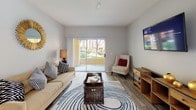 This single-floor residence offers a thoughtfully designed living space that balances comfort and style. The living area serves as the heart of the home, characterized by neutral gray walls and smooth, wood-look flooring that lends a contemporary yet warm aesthetic throughout. Large sliding glass doors flood the room with abundant natural light while framing a pleasant view of manicured greenery and a private patio, creating a seamless connection between the interior and the outdoors. The living space is generously proportioned, easily accommodating a full sofa ensemble, accent seating, and a central coffee table, all anchored by a patterned area rug that adds visual interest to the room. A wall-mounted flat-screen television is paired with a warm-toned media console, providing both functional and decorative appeal. Decorative touches, including a sculptural sunburst wall accent and accent pillows, contribute to an inviting, curated atmosphere. The overall ambiance is clean, bright, and modern, with a calm, neutral palette that makes the space feel open and airy. Natural light, outdoor access, and cohesive interior design come together to create a comfortable and practical living environment.