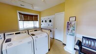 This single-floor shared laundry facility is designed to meet the practical needs of its users with a clean, functional layout. The room is brightened by warm yellow walls and natural light streaming through a window fitted with woven bamboo roller shades, creating a welcoming atmosphere. The flooring is a durable, easy-to-clean speckled vinyl, well-suited to a high-use environment. The space is outfitted with multiple full-size top-loading washing machines and dryers along one wall, complemented by a set of stacked washer-dryer units positioned against the back wall, maximizing capacity within the room. A countertop surface near the window provides a convenient area for folding or sorting laundry. Track lighting mounted on the ceiling ensures the space is well-lit throughout. A self-service payment and information kiosk is positioned near the entrance, offering easy access to machine operation details and payment options. A white interior door leads to an additional adjoining area. Overall, the facility is tidy, efficiently organized, and provides all the essentials for a comfortable and convenient laundry experience.