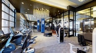 This premium fitness center is designed to deliver a high-end workout experience within an elegantly appointed space. The open floor plan is anchored by a stunning large-scale chandelier with cascading gold and crystal elements that serves as an eye-catching centerpiece, setting the tone for the refined aesthetic throughout.

Along one side of the facility, a row of modern cardio machines — including treadmills and ellipticals — lines the perimeter beneath large windows that allow natural light to pour in. Bold blue structural columns punctuate the space, adding a vivid contrast to the otherwise neutral palette of the carpeted flooring and dark ceiling treatment.

The functional training zone is well-equipped with free weights, resistance machines, and specialty equipment such as a heavy-duty tire for dynamic training exercises. A sleek glass-and-metal partition wall helps define separate workout areas while maintaining visual openness and depth across the floor. Full-height mirrors further amplify the sense of space.

Recessed lighting complements the chandelier, ensuring the entire facility is evenly and warmly illuminated. The overall ambiance balances the energy of a performance-driven gym with the sophistication of an upscale amenity space, making it a standout feature for residents or members.
