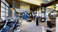 This premium fitness center is designed to deliver a high-end workout experience within an elegantly appointed space. The open floor plan is anchored by a stunning large-scale chandelier with cascading gold and crystal elements that serves as an eye-catching centerpiece, setting the tone for the refined aesthetic throughout.

Along one side of the facility, a row of modern cardio machines — including treadmills and ellipticals — lines the perimeter beneath large windows that allow natural light to pour in. Bold blue structural columns punctuate the space, adding a vivid contrast to the otherwise neutral palette of the carpeted flooring and dark ceiling treatment.

The functional training zone is well-equipped with free weights, resistance machines, and specialty equipment such as a heavy-duty tire for dynamic training exercises. A sleek glass-and-metal partition wall helps define separate workout areas while maintaining visual openness and depth across the floor. Full-height mirrors further amplify the sense of space.

Recessed lighting complements the chandelier, ensuring the entire facility is evenly and warmly illuminated. The overall ambiance balances the energy of a performance-driven gym with the sophistication of an upscale amenity space, making it a standout feature for residents or members.