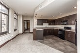 This single-floor urban residence offers a thoughtfully designed layout that balances open communal spaces with private retreats. The living area is expansive and filled with natural light, courtesy of a series of large, dark-framed windows that frame elevated city views of the surrounding urban landscape. Neutral carpet flooring runs throughout the living and bedroom areas, complementing the warm beige walls and rich dark wood baseboards.

The kitchen is well-appointed with dark espresso cabinetry extending to the ceiling, providing ample storage. Stone-toned countertops line the perimeter, and a peninsula island with additional cabinetry serves as a natural divider between the kitchen and living space. Black appliances, including a range, microwave, and refrigerator, complete the kitchen. Large-format tile flooring grounds the cooking area.

The bedrooms are generously sized, each featuring plush carpet, multiple windows with urban outlooks, and dedicated closet space. One bedroom includes a spacious walk-in closet with dual wire shelving systems, while another features a standard reach-in closet with hanging rod.

The bathrooms are finished with wood-look tile flooring and dark vanity cabinetry. One bathroom includes a tub-shower combination with sliding glass doors, while a second serves as a half or three-quarter bath with a streamlined vanity and toilet.