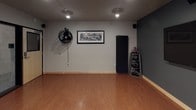 This single-floor dance and fitness studio is thoughtfully designed to support a wide range of movement-based activities. The main studio space is anchored by a full-length mirrored wall fitted with a classic wooden ballet barre, providing both functional support for dance training and the visual effect of an expanded, open space. The polished hardwood flooring throughout offers a smooth, resilient surface ideal for dance, yoga, and general fitness use.

The studio's walls are painted in a two-tone scheme — a calming neutral gray paired with a deeper charcoal accent wall — giving the space a modern, focused atmosphere. A large flat-panel display is mounted on the accent wall, enabling instructors or users to follow along with video content or display instructional media with ease. Track lighting mounted on the ceiling provides directed, adjustable illumination throughout the space.

Additional amenities include a wall-mounted circulation fan for climate control, a dedicated rack for kettlebells and fitness accessories, and a standing exercise mat for floor-based work. Near the entrance, a small service counter and a studio rules board contribute to the organized, professional feel of the space. The keypad-secured entry door ensures controlled access. The studio is compact yet highly functional, offering everything needed for a focused and versatile workout or training environment.