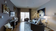 This single-floor residence showcases a thoughtfully designed living area that balances modern sophistication with comfortable functionality. The space is anchored by warm hardwood flooring that runs throughout, complemented by neutral-toned walls that create a versatile backdrop for the curated interior design.

The living room serves as the focal point of the space, featuring a generous seating arrangement with a light-toned sofa, dark accent chairs, and a curved sectional piece that defines the conversation area. A geometric-patterned area rug grounds the furniture grouping, adding texture and visual interest underfoot. A large-scale abstract artwork in navy and gold dominates one wall, contributing a bold artistic statement to the room.

Natural light floods the interior through expansive floor-to-ceiling sliding glass doors fitted with elegant floor-length drapery in a warm taupe tone. These doors open directly onto a private balcony with metal railings, offering views of the surrounding building exterior and landscaping. The balcony accommodates outdoor seating, extending the livable area beyond the interior.

Decorative wall accents in metallic gold add warmth along one wall, while a dark console table with styled accessories provides both function and visual depth. The overall ambiance is polished and contemporary.