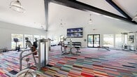 Located at 100 Cold Spring Rd in Rocky Hill, Connecticut, this fitness center offers a generously sized, single-floor layout designed to accommodate a comprehensive range of workout equipment and activities. The space is defined by its striking architectural character, including vaulted ceilings with exposed dark structural beams that add visual interest while contributing to an open, airy atmosphere.

A bold, multi-colored striped carpet covers the entire floor, lending the space an energetic and modern aesthetic. Large windows line multiple walls, flooding the interior with natural light and providing views of the surrounding outdoor amenities, including a pool area and landscaped grounds. Pendant lighting fixtures complement the natural light throughout the day.

The fitness center is equipped with cardio machines, including treadmills and ellipticals, a comprehensive free-weight area with a dumbbell rack, cable and strength machines, a TreadWall climbing machine, and rowing equipment. Full-length mirrors along one wall enhance the sense of space and support proper form during workouts. A wall-mounted multi-screen TV display is prominently positioned for entertainment. Entry is through sleek black-framed glass double doors, connecting to an adjacent facility.