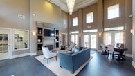 This elegantly designed single-floor lounge space immediately impresses with its dramatically soaring double-height ceilings, which lend an expansive, grand atmosphere throughout. A striking tiered crystal chandelier serves as the room's centerpiece, suspended high above and casting a warm, luminous glow across the interior. The walls are finished in a refined warm taupe, complemented by crisp white crown molding and architectural tray ceiling detailing that add depth and sophistication.

The seating area is thoughtfully arranged around a tufted navy velvet sofa paired with coordinating neutral accent chairs, all anchored by a light wood coffee table atop a textured area rug over rich dark hardwood flooring. A built-in media wall features an electric fireplace framed by open shelving with curated décor, adding both function and visual interest.

Natural light pours in generously through large, multi-paned windows and French doors positioned along the perimeter, offering views of an outdoor pool area and creating a seamless indoor-outdoor connection. Wall sconces provide ambient lighting throughout. The space exudes a polished, upscale ambiance ideal for gathering, relaxing, or socializing in comfort and style.