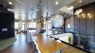 This single-floor space showcases a striking contemporary design defined by soaring ceilings and an open, expansive layout. At the heart of the main gathering area sits a substantial live-edge wood communal table surrounded by modern white chairs, serving as a natural focal point. Above, a dramatic cluster of cone-shaped pendant lights in dark matte finishes draws the eye upward, complementing the recessed lighting throughout the ceiling.

A glass-paneled railing lines a mezzanine-style overlook area, offering a sense of depth and visual continuity between levels. Beyond the railing, a lower section of the space features a dark granite countertop bar area, with a large landscape mural providing a scenic backdrop.

Natural light filters through multiple tall windows on multiple walls, brightening the interior and balancing the moody, sophisticated palette of dark ceilings and rich wood tones. A secondary lounge area with plush seating is visible in the background, adding versatility to the space. The overall ambiance is polished and upscale, blending industrial and modern design elements to create a refined and inviting communal environment.