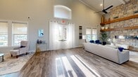 Located at 3 Vantage Way in Greenville, SC, this single-floor space makes a striking first impression with its soaring vaulted ceilings and grand double-door entrance framed by an arched transom window that floods the interior with natural light. Wide-plank wood-look flooring flows seamlessly throughout, anchoring the open, airy layout.

The expansive living area is a standout, dominated by a floor-to-ceiling stacked stone fireplace with a sleek linear burner and flanked by substantial rustic wood floating shelves. Large windows bathe the seating area in natural light, and a modern ceiling fan adds to the comfort of the space. The open layout transitions effortlessly into a dining area, where frosted-glass French doors lead to adjacent private rooms.

The kitchen is both functional and elegant, featuring crisp white cabinetry extending to the ceiling, a classic gray subway tile backsplash, stainless steel appliances, and a generously sized curved island topped with light granite countertops. Geometric chandelier pendants overhead add a contemporary design touch.

Together, the living and kitchen areas create a cohesive, welcoming environment defined by high-end finishes, architectural detail, and an abundance of natural light — ideal for both gathering and everyday use.