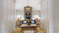 Located at 100 Riverchase Boulevard in Nashville, TN, this single-floor community space seamlessly combines a living room and dining area into one cohesive, well-appointed retreat. The interior is defined by soaring vaulted ceilings that draw the eye upward, anchored by a dramatic stacked stone fireplace serving as the room's focal point. A wrought-iron chandelier adds a refined yet approachable elegance to the space. Elegant arched doorways frame the living area, lending a classic architectural character reinforced by wainscoting and crisp white millwork throughout. Large French doors and generously sized windows flood the interior with natural light, offering views of the surrounding outdoor landscape and pool area. The living room features a warm, inviting arrangement with a plush sofa, accent chairs, and layered area rugs atop light tile flooring. A dedicated dining nook, situated near the French doors, offers a bistro-style seating area with high-top chairs, perfect for casual conversation. The neutral warm-toned palette of the walls pairs seamlessly with the natural textures of stone, wood, and woven materials, creating a balanced, welcoming ambiance throughout.