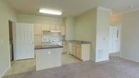 This single-floor space offers a practical and open layout that seamlessly blends the kitchen and living areas into one cohesive environment. The kitchen is well-appointed with light wood cabinetry that lines the walls, providing ample storage throughout. A center island with a dark countertop serves as a natural focal point, offering additional prep space and a visual anchor for the room. White appliances, including a range, microwave, and dishwasher, are neatly integrated into the cabinetry, while a double sink with a dedicated counter section rounds out the functional layout. The kitchen is finished with easy-to-maintain tile flooring, which transitions smoothly into soft, neutral carpeting that extends into the adjacent living area. Flush fluorescent lighting illuminates the kitchen, ensuring a bright and practical workspace. The living area benefits from the open sightlines into the kitchen, creating a connected and airy feel. Neutral wall tones throughout contribute to a clean, understated aesthetic that is both welcoming and versatile. A floor-level vent indicates a central heating system, and wall panels suggest thoughtful utility placement. Overall, the space presents a functional and tidy layout well-suited for everyday living.
