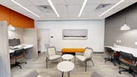 This contemporary workspace is designed to support both focused work and casual collaboration within a compact, efficiently utilized footprint. The space is defined by a striking color palette, featuring bold orange accent walls that contrast sharply with neutral white and dark gray surfaces, creating an energetic yet professional atmosphere throughout.

The open-plan layout is thoughtfully organized into two functional zones. A central lounge area features modern gray upholstered chairs arranged around round white side tables, complemented by a vibrant orange bench seat. A framed abstract artwork anchors the main white wall, adding a refined aesthetic touch. Adjacent to the lounge, a built-in workstation area offers multiple individual desk stations equipped with computer monitors, task chairs, and semi-private dividers, all illuminated by pendant dome lights overhead.

The flooring consists of patterned loop-pile carpet tiles in a neutral gray tone, providing both comfort and durability. A drop ceiling fitted with linear LED lighting panels ensures consistent, well-distributed illumination across the space. Large glass doors with black frames serve as the primary entry point, flooding the interior with natural light and offering views of the surrounding outdoor greenery. The overall design balances functionality with modern aesthetics, making it a productive and visually engaging environment.