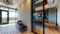 This refined single-floor space showcases a seamless blend of contemporary design and warm, luxurious materials throughout. The entry corridor immediately sets the tone with its light-toned brick accent wall, a bold geometric-patterned floor runner, and a statement chandelier overhead. Large floor-to-ceiling black-framed windows and a glass door at the corridor's end flood the space with natural light while framing sweeping panoramic views of the surrounding cityscape and greenery beyond a private balcony. A sleek leather bench provides an elegant seating moment along the corridor wall, complemented by a large-scale artwork on the opposing side. The open lounge area features coffered ceilings, wide-plank hardwood flooring, and an arrangement of plush, modern armchairs centered around a sculptural gold geometric coffee table. Built-in warm wood cabinetry with art glass sliding doors and an integrated bar niche with a backlit glass surface adds a bespoke architectural detail to this gathering space. The formal conference room is anchored by a live-edge wood table surrounded by rich teal velvet chairs, with floor-to-ceiling built-in shelving and cabinetry flanking a central wall-mounted display. Crystal chandeliers and recessed lighting throughout contribute to a polished, professional ambiance across the entire space.