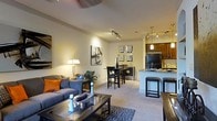 This single-floor residence features an open-concept layout that integrates the living, dining, and kitchen areas into one cohesive, flowing space. The living area is anchored by plush carpeted flooring, a comfortable sofa accented with warm orange and blue throw pillows, and a sleek glass-top coffee table. A patterned area rug adds texture and visual interest underfoot. Ambient lighting from a floor lamp and ceiling-mounted track lighting creates a warm, inviting atmosphere throughout the space. The dining area transitions naturally from the living room, offering a dark wood bistro-style table with matching chairs, ideal for casual meals. The kitchen is well-appointed with rich dark wood cabinetry, dark appliances, granite-style countertops, and pendant lighting hanging above a breakfast bar complete with two bar stools. Track lighting in the kitchen provides focused task illumination. Neutral-toned walls serve as a versatile backdrop, complemented by decorative wall art that adds personality to the space. The overall ambiance is warm, modern, and functional, making it well-suited for both relaxing and entertaining.