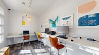 Located at 4106 Brickell Drive in Fairfax, VA, this single-room business center office is a thoughtfully designed workspace that balances functionality with contemporary aesthetics. The room features soaring vaulted ceilings and a generous wall of multi-pane windows along one side, flooding the interior with abundant natural light and offering views of the surrounding outdoor greenery. A track lighting system overhead supplements natural illumination throughout the day.

The workspace is anchored by continuous white built-in countertop desks that run along multiple walls, providing ample surface area for several workstations equipped with desktop computers, a printer, and shared peripherals. Warm-toned wooden chairs with a modern silhouette line the desks, adding a stylish contrast to the crisp white surfaces and walls. A natural wood bench positioned beneath the windows provides additional seating.

The floor features a patterned, textured carpet in cool grey tones, lending visual interest to the space. The walls are adorned with a curated selection of framed abstract artworks in vibrant colors—including blues, teals, and warm yellows—enhancing the creative and professional ambiance. A glass-paned entry door connects the office to the broader facility. Overall, the space presents a clean, bright, and productive environment well-suited for focused work.