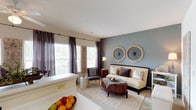 The Edgartown is a single-story, 3-bedroom, 2-bathroom residence designed with a cohesive, transitional aesthetic throughout. The living room serves as the centerpiece of the home, featuring a distinctive blue-gray accent wall adorned with circular medallion artwork, neutral carpet flooring, and multiple windows dressed with floor-length curtains that allow ample natural light. The space flows directly into the kitchen, which features a U-shaped layout with white cabinetry accented by warm wood trim, white appliances, light countertops, and a peninsula with a built-in sink that opens toward the living area. Each of the three bedrooms is carpeted and includes windows for natural light and closet access. The bedrooms vary in scale, with the largest featuring a ceiling fan and decorative shutter wall art, while the additional bedrooms offer cozy, well-proportioned spaces with their own windows and closets. The two bathrooms include tile flooring, vanities with wide mirrors, and shower-tub combinations. Throughout, the residence maintains a warm, neutral palette with consistent design details, creating a comfortable and functional living environment.