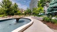 Located at 17 Lascelles Blvd in the prestigious Forest Hill South neighbourhood of Toronto, this outdoor amenity space offers a thoughtfully designed communal grounds that serves as a verdant retreat within an urban residential setting. At its centerpiece sits a generously sized freeform pool featuring multiple water fountain elements, its organic curved shape complemented by a smooth stone surround that invites leisurely strolling and relaxation. The water features create a calming ambiance, with gentle jets and a central fountain adding both visual interest and the soothing sound of flowing water. Surrounding the pool, the landscape is richly planted with a diverse mix of mature deciduous trees, blue spruce, ornamental shrubs, and layered perennial gardens that provide lush, year-round texture and colour. Paved walkways wind throughout the grounds, connecting various seating areas furnished with benches and framed by manicured plantings and natural stone accents. Elevated deck structures with white railings add architectural dimension to the landscape, accessible via sweeping staircases. Globe-style lamp posts line the pathways, providing both a classic aesthetic touch and practical illumination. The entire courtyard is framed by the surrounding residential towers, creating an intimate, park-like setting that offers residents a peaceful outdoor escape in the heart of midtown Toronto.
