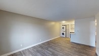 This single-floor space features a clean, open layout with a neutral color palette of soft grey walls and white baseboards, creating a bright and modern atmosphere. The main living area is generously proportioned, with durable laminate wood-look flooring that extends throughout, offering both style and easy maintenance. A textured ceiling adds subtle character to the space.

Toward the rear of the unit, a functional in-suite laundry area houses a stacked washer and dryer unit, conveniently positioned alongside a full-size refrigerator. Adjacent to the laundry area, the kitchen is partially visible and features white cabinetry, providing ample storage, as well as a countertop that transitions into a breakfast bar or pass-through-style overhang — ideal for casual dining or additional prep space. The kitchen benefits from its own dedicated lighting, keeping the area well-illuminated.

Overall, the space offers a practical and well-organized layout suited for everyday living, with modern finishes and thoughtful amenities integrated throughout.