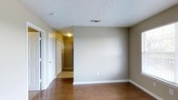 This single-floor space presents a clean, straightforward layout designed for functional living. The main room is finished with rich, dark hardwood flooring that adds warmth and depth to the interior, complemented by neutral greige-toned walls that provide a versatile backdrop for a variety of décor styles. The textured popcorn ceiling features a central HVAC vent, ensuring efficient climate control throughout the space.

A doorway in the corner opens to a connecting hallway, which provides access to additional areas of the unit. Visible from the main room, the hallway is illuminated by a flush-mount ceiling fixture and lined with bifold closet doors, offering practical storage solutions. The hallway flooring transitions to tile, adding a subtle contrast to the hardwood found in the primary room.

White trim and baseboards run throughout, providing a crisp, defined border between the walls and floors. The space benefits from natural light entering through what appears to be a sliding glass door or large window on the left side of the room. Overall, the space offers a neutral, move-in-ready environment with functional storage and a cohesive, understated aesthetic.
