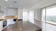 This single-floor space showcases a sophisticated open-concept design that seamlessly integrates the kitchen and living areas into one expansive, luminous environment. Floor-to-ceiling glass windows and sliding doors line the living area, flooding the interior with abundant natural light while framing sweeping panoramic views of the surrounding cityscape and sky beyond.

The modern kitchen is appointed with crisp white cabinetry, a warm gold-toned tile backsplash, and a generous stone countertop island that anchors the space. A professional-style pull-down faucet and an undermount sink add functional elegance, while built-in appliances maintain the kitchen's clean, streamlined aesthetic.

The open living area extends naturally from the kitchen, featuring warm-toned hardwood flooring that runs throughout and a ceiling fan that enhances the airy, comfortable atmosphere. The bright white walls and smooth ceilings reflect natural light throughout the day, contributing to the space's fresh, contemporary feel. The seamless flow between the kitchen and living zones makes this an ideal setting for both everyday living and entertaining, defined by clean lines, high-end finishes, and a sense of openness rarely found in urban settings.