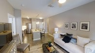 This well-appointed single-floor residence showcases a thoughtfully designed open-concept layout that seamlessly integrates the kitchen, dining, and living areas into one cohesive and functional space. The kitchen is a standout feature, boasting stainless steel appliances, sleek cabinetry in a warm gray-taupe finish, and a central island with bar seating — ideal for casual dining or entertaining. Pendant lighting above the island adds an elegant touch, complementing the recessed lighting throughout. The flooring features wood-look tile laid in a linear pattern, lending warmth and continuity across the entire space. The living area is comfortably proportioned, anchored by a neutral-toned sofa accented with dark decorative pillows, a tufted ottoman serving as a coffee table, and a classic table lamp that adds ambient warmth. Soft, neutral wall tones in beige and greige create a calming and versatile backdrop throughout. A ceiling fan in the living area enhances comfort, while framed wall art adds a curated, refined aesthetic. The overall ambiance is modern yet inviting, with clean lines and a cohesive color palette that makes the space feel both polished and livable.
