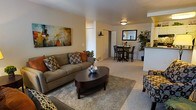 This single-floor residence offers a thoughtfully arranged open-concept floor plan that seamlessly connects the living and dining areas, creating a cohesive and functional living environment. The living room is anchored by a plush, neutral-toned sofa accented with decorative throw pillows in warm earth tones, complemented by an oval dark-wood coffee table and a textured area rug. A bold abstract canvas artwork adds a pop of color to the neutral walls, enhancing the room's inviting ambiance. Soft carpeting throughout the main living space adds warmth and comfort underfoot. The dining area, visible from the living room, features a dark bistro-style table with matching chairs, ideal for intimate gatherings. A large rectangular mirror on the dining room wall reflects light and adds a sense of depth to the space. Adjacent to the dining area, a partial view of the kitchen reveals a peninsula counter with bar-height seating, hinting at a practical and connected culinary space. A tall indoor plant near the kitchen adds a touch of greenery, while the overall neutral color palette and warm lighting create a calm, comfortable atmosphere throughout this well-designed home.