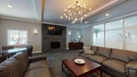 Located at 1 Juniper in Yarmouth, Maine, this single-floor community lounge is a thoughtfully designed gathering space that blends comfort with contemporary style. The centerpiece of the main lounge area is a floor-to-ceiling stacked stone fireplace, topped with a wall-mounted television and flanked by subtle sconce lighting, creating a warm and inviting focal point. Overhead, a striking sculptural chandelier with an branching metallic frame adds a dramatic decorative element to the tray ceiling, which is further enhanced by recessed lighting throughout.

The lounge accommodates multiple seating configurations, including plush sofas and accent chairs arranged around a rich wood coffee table, as well as a separate dining area with a round table and upholstered chairs — ideal for casual meetings or social gatherings. Soft, patterned carpet in muted tones runs throughout the space, complementing the cool gray wall palette.

A wall of frosted glass panels along one side of the room adds architectural interest while allowing diffused light to filter through. Sliding glass doors open to the exterior, filling the space with natural light and outdoor views. A compact kitchenette equipped with dark countertops, white cabinetry, a stainless steel sink, microwave, and full-size refrigerator provides practical convenience for residents and guests alike.