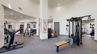 The Los Alisos at Mission Viejo Fitness Center is a 2,340-square-foot, single-floor facility designed to support a comprehensive workout experience. The space features a striking architectural design with soaring ceilings, curved walls, and an abundance of recessed lighting that creates a bright, energizing atmosphere. High clerestory windows allow natural light to pour into the main exercise area, enhancing the open and airy feel throughout.

The fitness floor is divided into distinct functional zones. The cardio area is outfitted with Life Fitness treadmills, elliptical trainers, upright bikes, and recumbent cycles, all positioned on hardwood-style flooring. The strength training zone features a multi-station cable machine, free-weight benches, and a comprehensive dumbbell rack with weights displayed along mirrored walls that amplify the sense of space. A dedicated functional training area includes kettlebells, stability balls, and additional benches on durable rubberized flooring.

Large black-framed glass partition doors separate sections of the facility, while curated abstract wall art adds a polished, modern aesthetic. Wall-mounted flat-screen TVs are positioned near cardio equipment for entertainment. A wall-mounted water station with a bottle-filling dispenser ensures convenient hydration. The seamless blend of rubberized and hardwood flooring zones defines each workout area while maintaining a cohesive, contemporary design throughout.