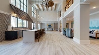 This expansive single-floor amenity space is designed with a coastal-modern aesthetic, blending sophisticated architectural details with relaxed, inviting interiors. The centerpiece of the space is a dramatic great room boasting soaring double-height ceilings, exposed wood beam accents, and a striking oversized windmill-style ceiling fan. Floor-to-ceiling windows and clerestory glazing flood the area with abundant natural light, while black-framed French doors open to an outdoor terrace beyond.

Light-toned wood-look flooring spans much of the space, transitioning to deeper wood tones in the lounge and dining zones, adding visual depth and definition to each area. A wet bar with a long white countertop, bar seating, a decorative metal canopy accent, and patterned tile backsplash serves as a focal gathering point. Nearby, a charming rope-hung porch swing provides a unique and playful seating option alongside café-style dining tables and bar-height seating.

The lounge area features plush leather sofas, accent chairs, a built-in media wall, and bold geometric wallcoverings. Woven pendant lights illuminate a high-top communal table, while soft aqua accent walls and coastal artwork carry a breezy, resort-inspired palette throughout the corridors and secondary rooms, creating a cohesive and welcoming atmosphere.