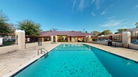 Located at 9801 Meadowglen Ln in Houston, Texas, this outdoor amenity area centers around a generously sized swimming pool with vivid turquoise water, ideal for recreation and relaxation. The pool is framed by a broad, light-colored concrete deck that provides ample space for lounging and movement around the water's edge. Entry into the pool is facilitated by stainless steel handrails and built-in steps, offering convenient and safe access. A distinctive single-story clubhouse building with a signature burgundy metal roof sits adjacent to the pool, featuring warm earth-toned exterior walls and arched architectural accents that lend a welcoming character to the space. The facility is enclosed by a sturdy wrought iron perimeter fence with structured masonry columns, ensuring both security and a defined boundary for the amenity area. Mature trees line the outer edges of the property, providing natural shade and a green backdrop against the open Texas sky. A covered gazebo structure is also visible within the fenced area, offering additional shelter for outdoor gatherings. The overall setting is well-maintained and open, benefiting from abundant natural sunlight and a clear view of the surrounding residential community.