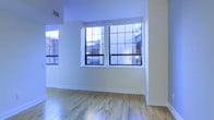 This two-floor space combines clean, modern finishes with subtle industrial character. The interior is defined by smooth white walls and light-toned hardwood floors that run continuously, lending a bright and cohesive feel to the space. Prominently featured are large, multi-pane windows with bold black frames — a nod to industrial loft-style architecture — which allow generous amounts of natural light to filter in while framing views of the surrounding urban environment. The windows are positioned to maximize daylight penetration, keeping the interior feeling open and airy even without additional lighting. A ceiling-mounted ventilation vent is integrated cleanly into the upper wall, reflecting thoughtful infrastructure detailing. The white walls provide a neutral, versatile backdrop suitable for a variety of uses or configurations. The overall ambiance is minimalist and functional, with an understated aesthetic that balances contemporary finishes with classic loft-inspired architectural elements. The two-floor layout offers flexibility and separation of space, making it well-suited for both living and working arrangements. The light, uncluttered interior creates a calm and adaptable environment throughout.