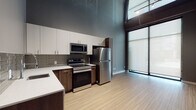 This contemporary single-floor space showcases a striking modern design defined by dramatic double-height ceilings and an open, airy layout. The kitchen is a standout feature, outfitted with a full suite of stainless steel appliances including a refrigerator, electric range, microwave, and dishwasher. Cabinetry combines crisp white upper cabinets with rich dark wood lower cabinets, complemented by a clean white countertop and a tasteful horizontal tile backsplash in muted earth tones. Light bamboo-style hardwood flooring flows throughout, adding warmth to the space's cool gray palette. A dramatic floor-to-ceiling window wall floods the kitchen and living area with abundant natural light, creating a bright and welcoming atmosphere. An additional room with neutral gray walls and the same light hardwood flooring offers flexible use as a bedroom or office. The bathroom is elegantly appointed with large-format marble-look wall tiles extending from floor to ceiling, a rich dark wood vanity, white countertop, and a backlit LED mirror that adds a refined, modern touch. Together, these elements create a cohesive, upscale urban living environment with a sleek industrial-modern aesthetic.