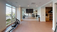 Located in the heart of Tampa at 111 W Fortune St, this contemporary fitness center offers a well-appointed, single-floor workout facility designed with both function and aesthetics in mind. The space is bathed in natural light, courtesy of expansive floor-to-ceiling windows that frame views of the surrounding greenery and streetscape, creating an energizing atmosphere for exercise. The clean white walls are complemented by a standout textured accent wall composed of layered stone and reclaimed wood elements, adding a bold artistic focal point throughout the space. A large wall-mounted flat-screen television anchors one end of the room, reflected in a full-length mirror wall that amplifies the sense of depth and openness. The facility is comprehensively equipped with a dedicated cycling studio featuring multiple stationary bikes, rowing machines by Matrix, a free weight station with a full dumbbell rack, and additional strength and cardio equipment. Glass partitions elegantly divide workout zones while maintaining visual connectivity across the floor. Durable, easy-clean flooring in a warm neutral tone runs throughout, and recessed lighting ensures consistent illumination across all areas. The overall design strikes a balance between modern sophistication and practical fitness functionality.