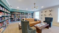 This thoughtfully designed library and lounge space combines functionality with refined aesthetics across a single level. The most striking feature is the expansive floor-to-ceiling built-in bookshelf wall, painted in a bold teal hue and filled with a wide-ranging collection of books, creating a warm and intellectual atmosphere throughout the room.

The lounge area is anchored by a cognac leather sofa paired with plush teal velvet accent chairs and a wooden coffee table, all centered beneath a mid-century modern brass and frosted glass chandelier. Recessed lighting and crown molding add a polished architectural detail to the ceiling. Light wood-look flooring runs through this section of the space, contributing to a bright, welcoming feel.

A separate activity zone features a long dark wood table surrounded by upholstered dining chairs, positioned alongside additional teal bookshelves and a large window that invites natural light. An adjacent workstation area offers two computer desks with ergonomic chairs, built-in cabinetry, and ample counter space. Decorative air plant wall art displayed on soft neutral walls adds a unique, nature-inspired design element throughout the space.