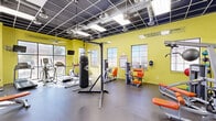 This fitness center offers a generously sized, single-floor workout space designed to support a wide range of exercise routines. The room is immediately distinguished by its bold yellow-green accent walls, which lend an energetic and motivating atmosphere throughout. Inspirational quotes are displayed along the upper walls, reinforcing a focus on fitness and perseverance.

The space is outfitted with a comprehensive mix of equipment, including treadmills, elliptical machines, and a stationary bike for cardiovascular training, alongside a variety of strength machines featuring striking orange upholstery. A TKO heavy punching bag and freestanding bag rack provide options for combat-style training, while a dumbbell rack and weight bench accommodate free weight workouts. A pull-up and dip station adds further versatility.

Large multi-pane windows line the walls, flooding the space with natural light and offering views to the surrounding outdoor areas. The dark drop ceiling with exposed ductwork and grid-mounted fluorescent lighting creates an industrial aesthetic that contrasts effectively with the bright walls. The flooring consists of durable rubber gym matting, providing safety and comfort underfoot. A wall-mounted television and large mirrors further enhance the functionality and feel of this well-appointed fitness facility.