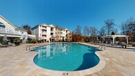 Located at 655 W Mallard Creek Church Rd in Charlotte, NC, this outdoor amenity space is centered around a generously sized, freeform resort-style swimming pool with clear blue water and a classic stone-tiled surround. Multiple entry points with stainless steel handrails provide easy pool access, while a broad deck lined with lounge chairs offers ample space for sunbathing and relaxation under the open sky.

Adjacent to the pool, a covered outdoor lounge area provides a shaded retreat equipped with cushioned seating, ceiling fans, and a tiled floor, making it comfortable even on warm Carolina days. This covered pavilion connects directly to a well-maintained restroom and changing facility block, which features multiple gender-designated rooms clearly marked with accessibility signage, a water fountain station, and clean concrete flooring beneath a beadboard-paneled ceiling.

The entire amenity area is enclosed by ornamental iron fencing with a gated entry, ensuring both security and a polished aesthetic. Manicured landscaping and mature trees frame the surroundings, with multi-story residential buildings visible in the background. The space benefits from abundant natural light and an open, airy atmosphere, making it a functional and inviting community gathering destination.