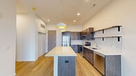 This single-floor residence showcases a clean, contemporary design with a thoughtful layout across its living spaces. The kitchen serves as a central highlight, featuring rich dark-toned cabinetry, a full suite of stainless steel appliances including a refrigerator, range, microwave, and dishwasher, and a classic white subway tile backsplash. A generously sized island with a crisp white countertop anchors the space, illuminated by a pair of stylish pendant lights with gold-accented interiors. Light hardwood-style flooring flows throughout the kitchen and living areas, adding warmth to the modern palette.

The bedrooms are finished with plush neutral carpeting and feature large windows that invite natural light and offer open outdoor views of the surrounding landscape. One bedroom includes sliding glass doors leading to a private balcony with expansive exterior views, further enhancing the sense of openness.

Two bathrooms serve the unit. The first is a full bathroom equipped with a combined shower and soaking tub, a dark wood vanity with a white undermount sink, and a large mirror. The second is a three-quarter bathroom with a standalone walk-in shower, toilet, and vanity. Both bathrooms maintain the unit's clean, minimalist aesthetic with light flooring and crisp white fixtures throughout.