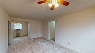 This single-floor unit offers a straightforward, functional layout across several rooms, as visible in the 3D floor plan overview. The living and bedroom spaces are finished with neutral-toned wall-to-wall carpeting and painted in soft, off-white tones, creating a clean and understated aesthetic throughout. Each room is equipped with a ceiling fan fitted with a built-in light fixture, providing both air circulation and overhead lighting. Several rooms feature classic cast-iron radiators beneath windows fitted with horizontal blinds, indicating a radiant heating system. One room is additionally equipped with a window-mounted air conditioning unit. Closet space is present in multiple rooms, including at least one walk-in style closet. The bathroom retains a classic design with white subway-style wall tiling accented by a bold navy border, a pedestal sink, a mirror, and a tub-shower combination with a window providing natural light. The kitchen includes light maple cabinetry, a white countertop, and a stainless steel double-basin sink. A chandelier accent light is present in one of the bedrooms, adding a decorative touch to the otherwise simply appointed space.